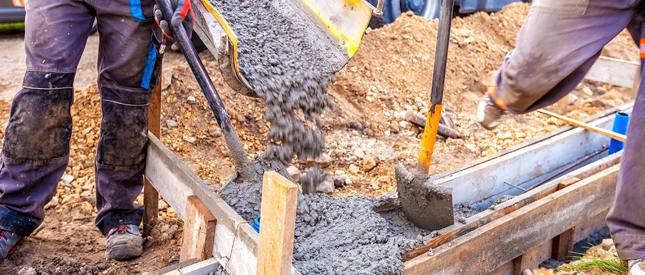 Zwei Bauarbeiter gießen Beton in eine vorbereitete Schalung, um das Fundament für eine Terrassenüberdachung zu schaffen. Werkzeuge und Baufahrzeug im Hintergrund zeigen den aktiven Montageprozess.