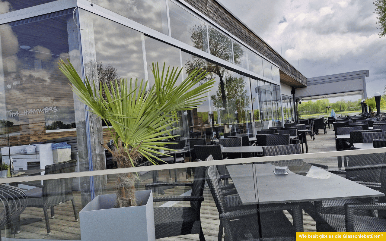 Wie breit gibt es die Glasschiebetüren? Moderne Glasfassade mit Schiebetüren auf einer Terrasse vor dem Restaurant mit Sitzplätzen und Pflanzen.