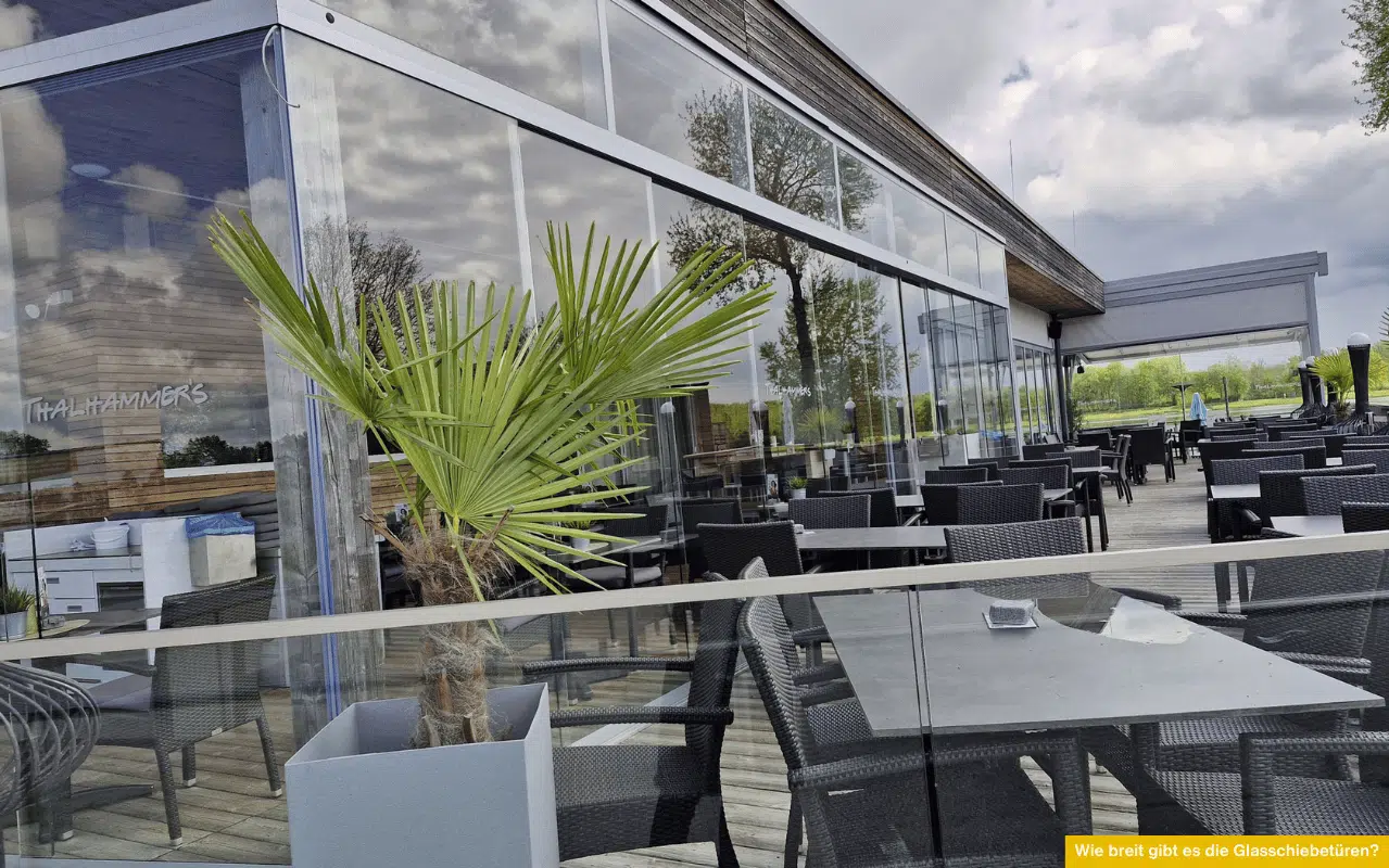 Wie breit gibt es die Glasschiebetüren? Moderne Glasfassade mit Schiebetüren auf einer Terrasse vor dem Restaurant mit Sitzplätzen und Pflanzen.