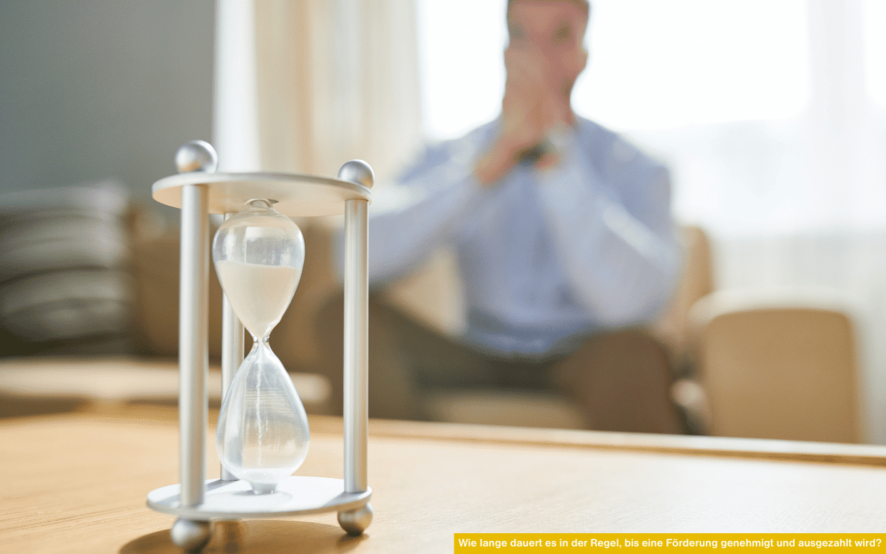Eine Sanduhr mit herabrieselndem Sand symbolisiert das Warten. Dahinter sitzt verschwommen ein Mann auf einem Sessel.
