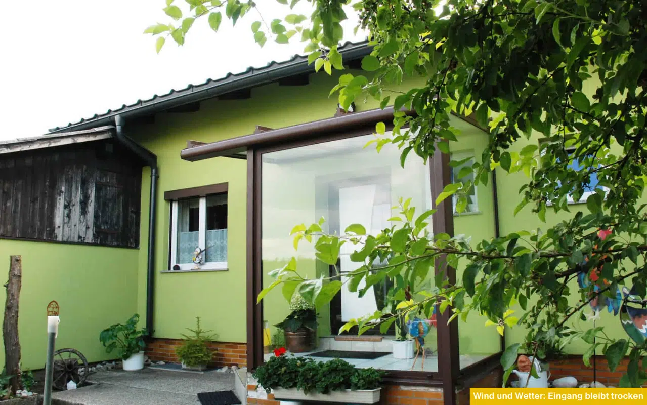 Wind und Wetter Eingang bleibt trocken moderner verglaster Windfang vor grünem Haus schützt den Eingangsbereich vor Regen und Feuchtigkeit.