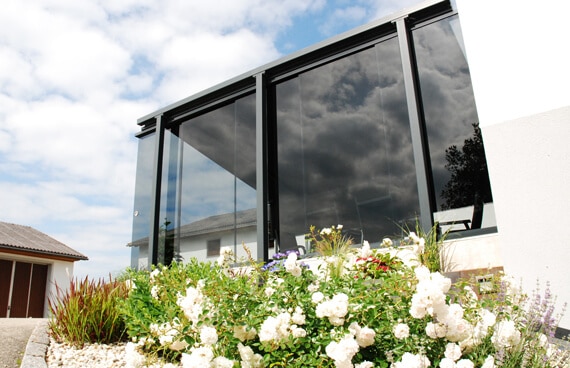 Terrassenbereich mit hoher Glaswand, die als Windschutz dient. Vor dem Haus ein bepflanztes Beet mit weißen und grünen Pflanzen, darüber blauer Himmel mit Wolken.