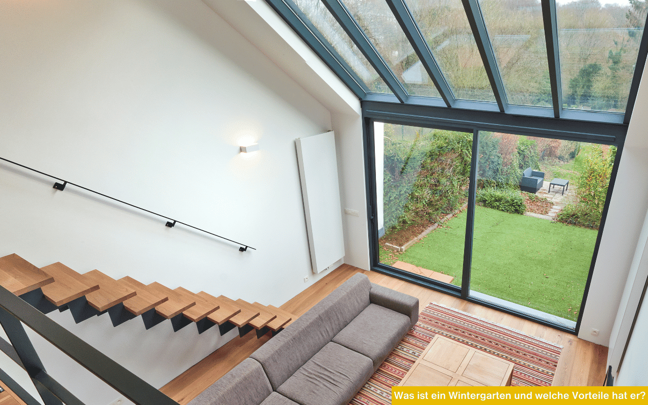 Blick von oben in einen modernen Wohnraum mit großer Glasfront, Holztreppe und Sofa, mit Aussicht auf einen grünen Garten.