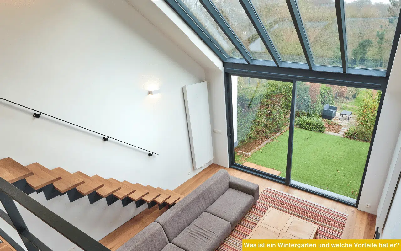 Blick von oben in einen modernen Wohnraum mit großer Glasfront, Holztreppe und Sofa, mit Aussicht auf einen grünen Garten.