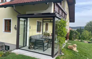 Ein Hund liegt entspannt auf dem Rasen neben einem verglasten Sommergarten, in dem eine Katze auf dem Boden schläft.