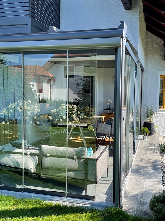 Moderner Wintergarten mit bodentiefen Glasschiebetüren. Innen stehen Sofa und Tisch, außen Garten und Terrasse. Die Glasflächen lassen viel Licht herein und verbinden Innen- und Außenbereich.