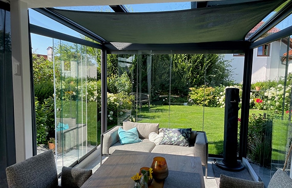 Ein moderner Wintergarten mit Glasschiebetüren, Sitzmöbeln und Tisch. Durch die transparenten Glasflächen ist ein gepflegter Garten mit Rasen und Pflanzen sichtbar. Viel Tageslicht sorgt für eine offene, wohnliche Atmosphäre.