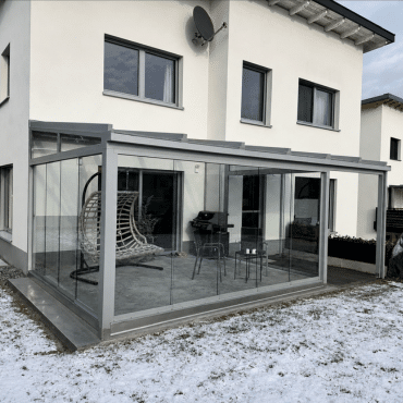 Wintergarten in Freistadt mit grauem Aluminiumrahmen und Ganzglas-Schiebeelementen an einem weißen Wohnhaus im Winter