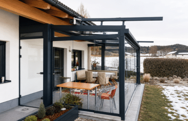 Ein moderner Wintergarten in Perg mit dunklem Aluminiumrahmen und großen Glasflächen erweitert die Terrasse eines Einfamilienhauses. Im Inneren stehen Esstisch und Stühle, im Hintergrund sind Garten und winterliche Landschaft zu sehen.