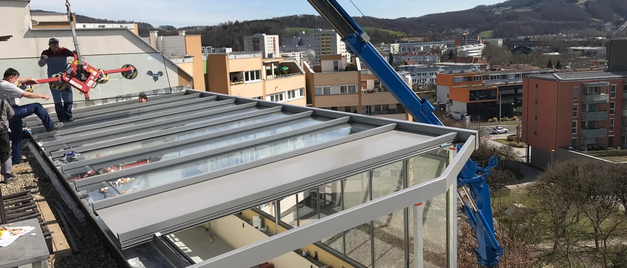 Auf einer Dachterrasse wird ein Wintergarten montiert. Ein Kran mit Sauggreifer hebt Glaselemente bzw. Dachprofile, daneben arbeiten Monteure am Aluminiumrahmen. Im Hintergrund sieht man Linz und umliegende Hügel.