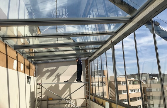 Innenaufnahme einer Wintergartenmontage: Ein Arbeiter justiert die Dachverglasung auf einem Gerüst. Der Raum ist hell, großflächig verglast und zeigt den Blick auf die Stadt Linz im Hintergrund.