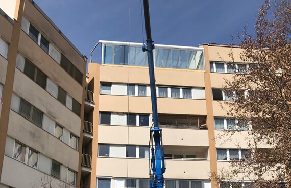 Vor einem mehrstöckigen Wohnhaus steht ein großer blauer Kran. Er hebt Glaselemente für den neuen Wintergarten auf das Dach der obersten Etage. Der Wintergarten ist teilweise bereits aufgebaut und verglast.