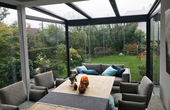 Modernen Wintergarten mit rustikalem Holztisch, grauen Polsterstühlen und Blick ins Grüne. Kerzen und Kissen schaffen eine warme, wohnliche Atmosphäre – perfekt für entspannte Stunden zu jeder Jahreszeit.
