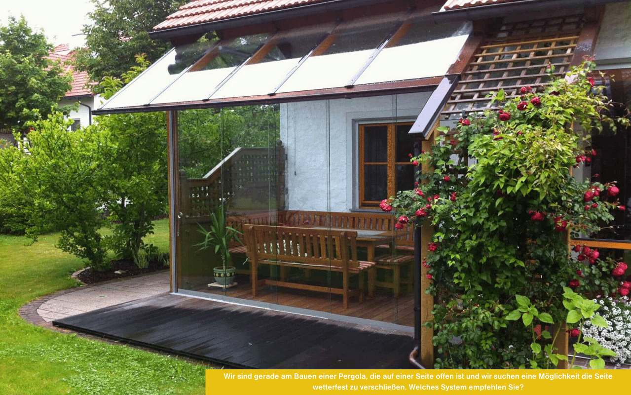 Gartenpergola am Haus mit teilweise verglaster Seite. Sitzbank und Tisch stehen geschützt, daneben Pflanzen und rote Kletterrosen.