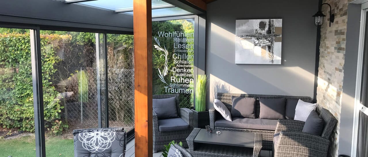 Ein moderner Wintergarten mit Glasdach, Holzbalken und großer Glasfront. Darin stehen graue Lounge-Möbel, ein Tisch und Deko. Der Raum wirkt offen, hell und einladend.
