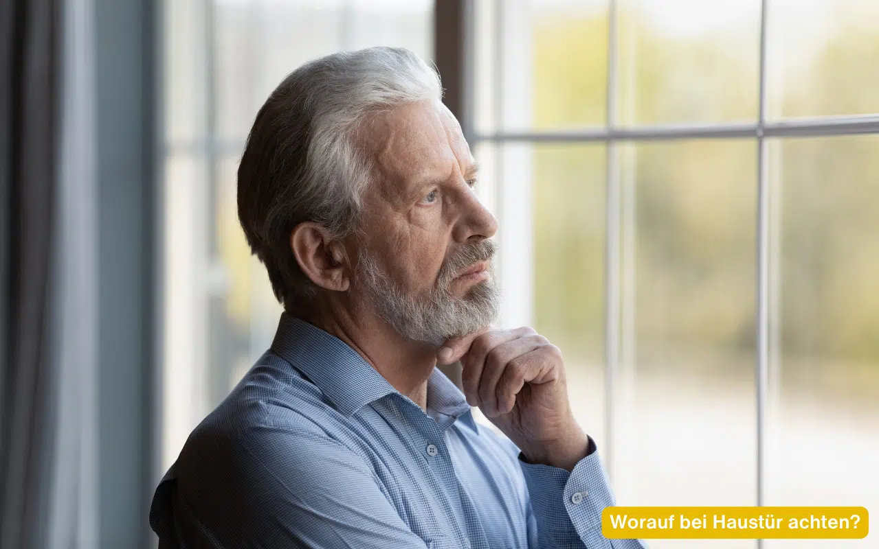 Worauf bei Haustür achten? Ein älterer Mann mit grauem Bart blickt nachdenklich aus einem Fenster mit Sprossen